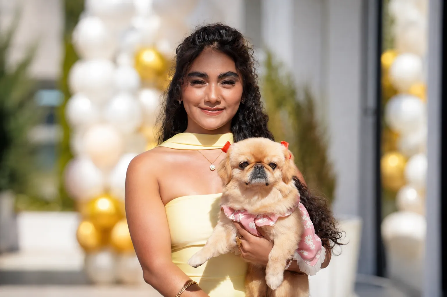 Retrato de una invitada sonriendo con su perro pequinés en brazos, entre columnas de globos dorados y blancos